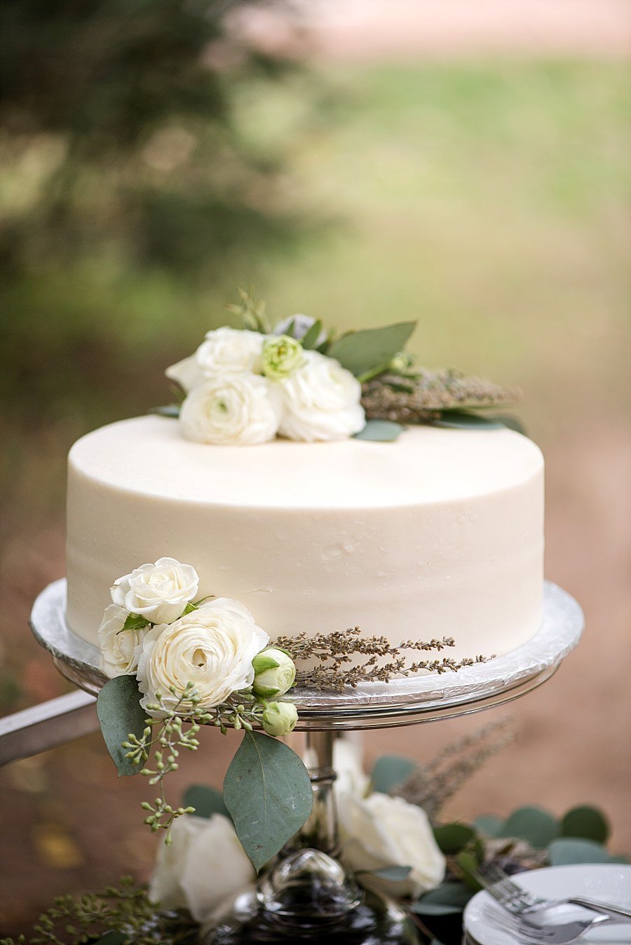The low one tier wedding cake with white icing sits on a silver pedestal cake stand decorated with white ranunculus and greenery.