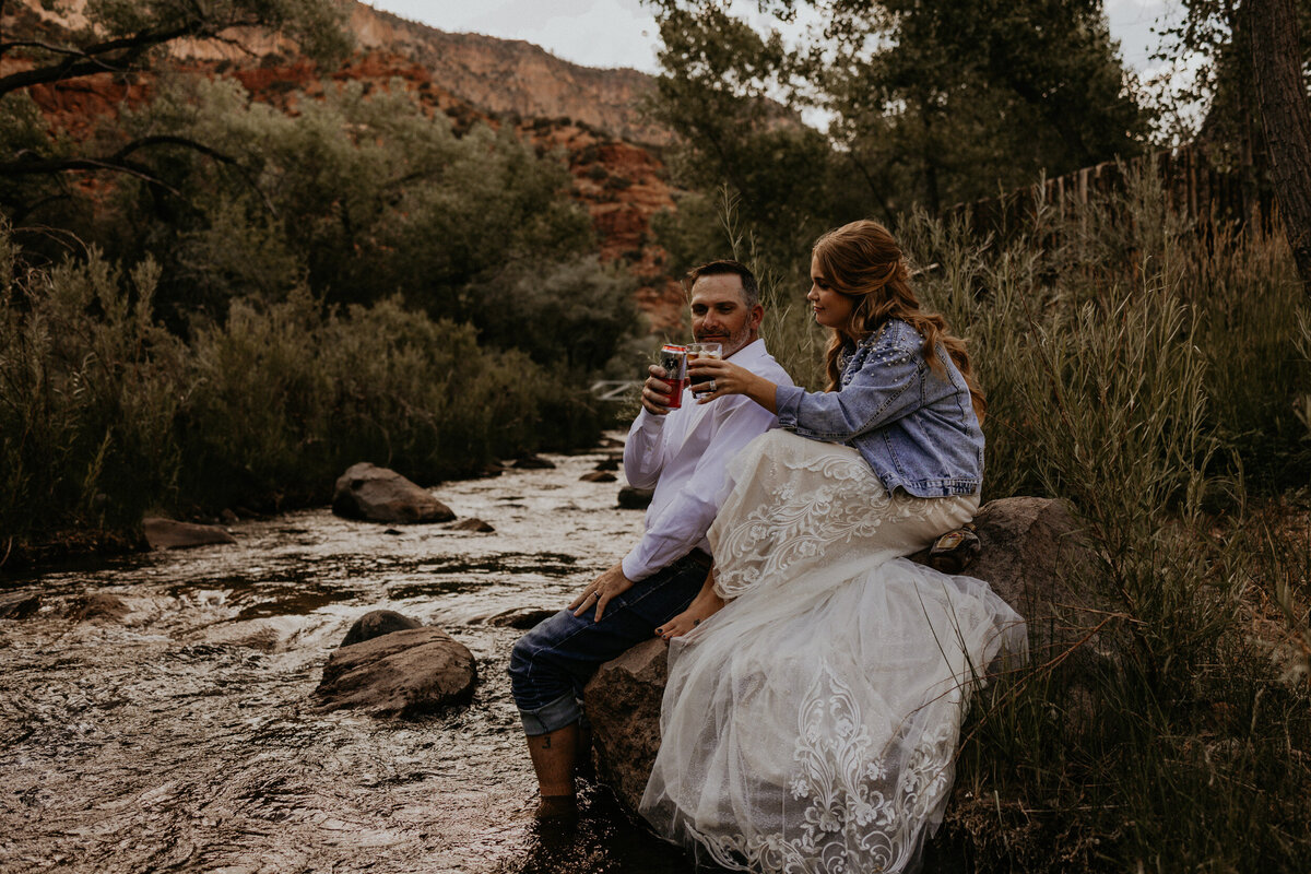newlyweds enjoying a drink together sitting along a creek after their  elopement