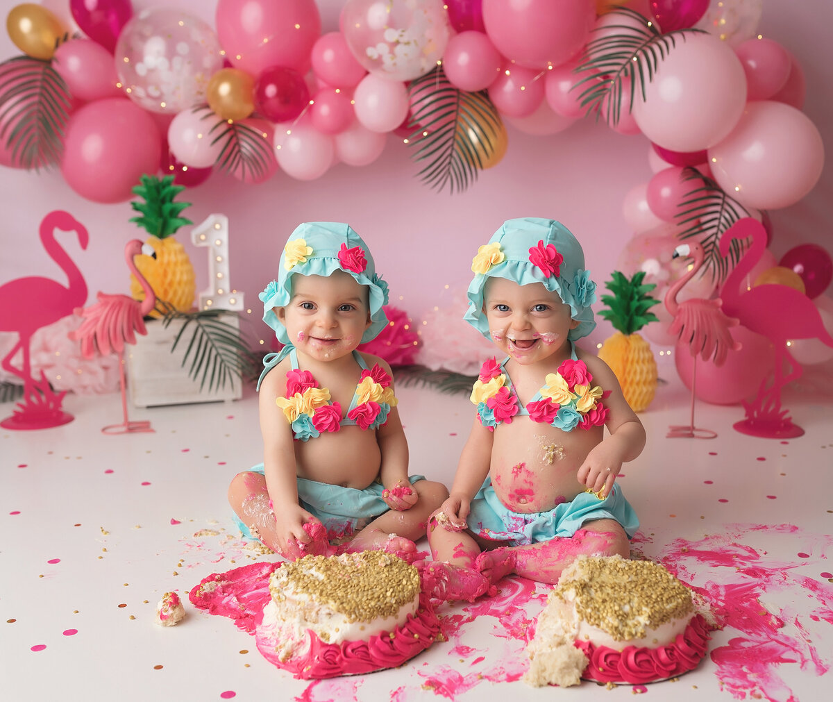 Hawaiian flamingo themed cake smash in West Palm Beach photography studio. Twin baby girls are in pink, yellow, and teal floral bikini tops with matching hats and teal skirts. They are smiling at the camera with the cakes smashed in front of them. In the background, there is a balloon arch with various shades of pink and interwoven leaves, pineapples, and flamingos.