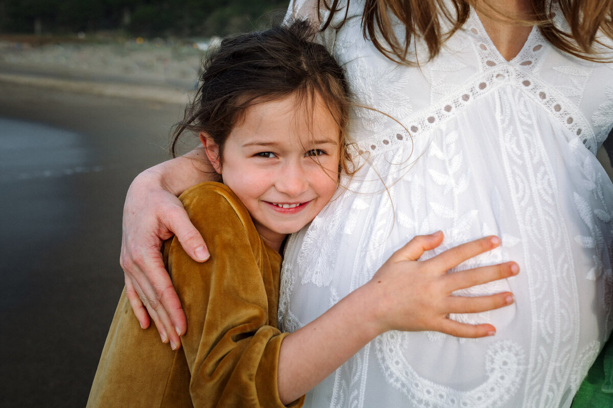 7-year-old girl hugging mom's baby bump and smiling at the camera at San Francisco beach