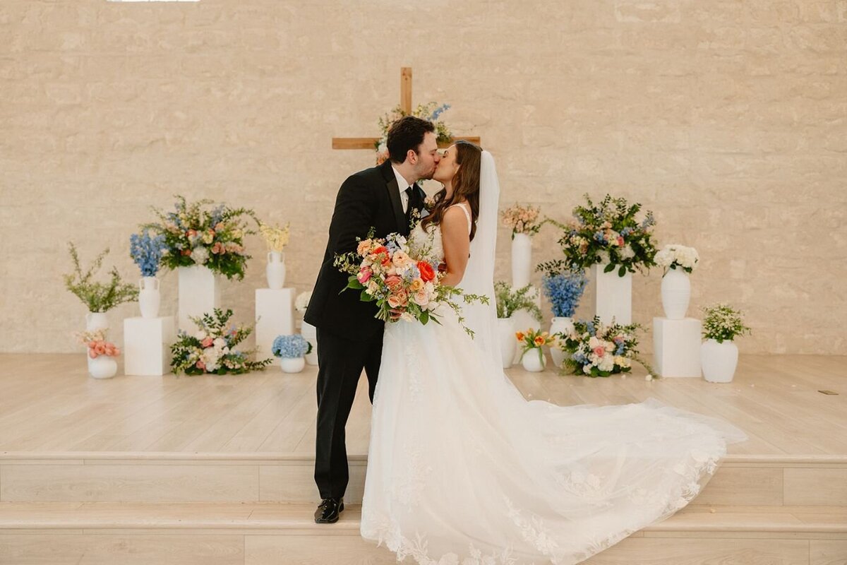 Bride and groom kiss at chapel altar surrounded by colorful floral arrangements with vibrant blooms and blue accents