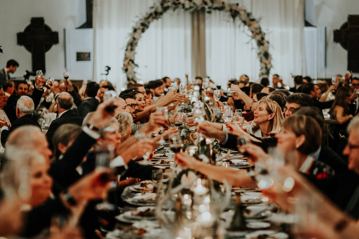 A very long harvest table with many guests happily clinking glasses at the end of a wedding toast with a large focal floral moongate arch at the end of the room at AllSaints venue, a converted church  in Ottawa Ontario