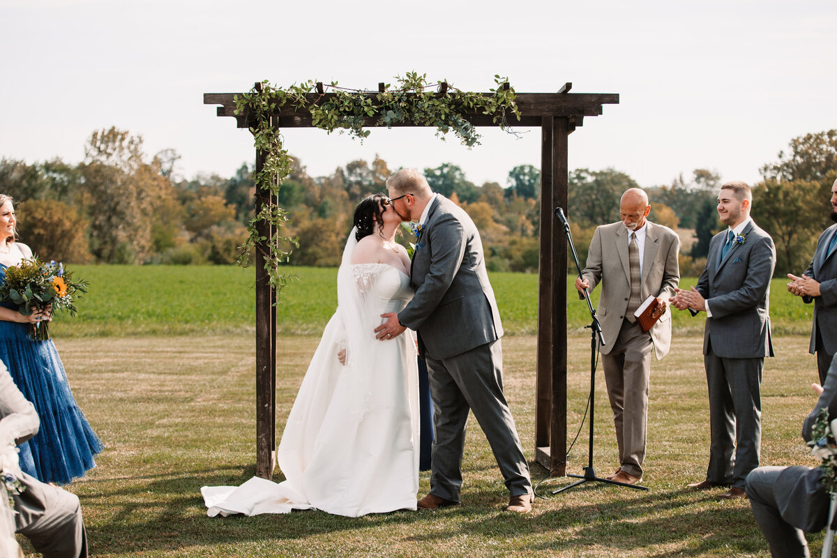NEwlyweds kiss under a wooden arbor to end their ceremony