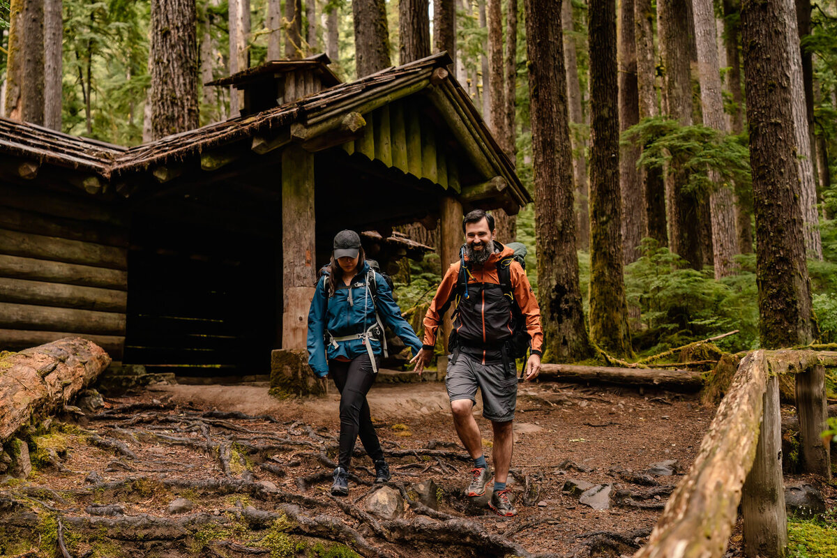A couple ends their Olympic National Park Elopement by changing into their hiking gear to set out on another adventure. they walk away from a log cabin covered in moss, nestled in the forest.