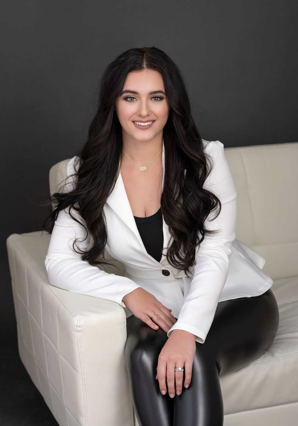 A woman in a cream blazer and fitted pants, leaning on a velvet couch, exuding professionalism.