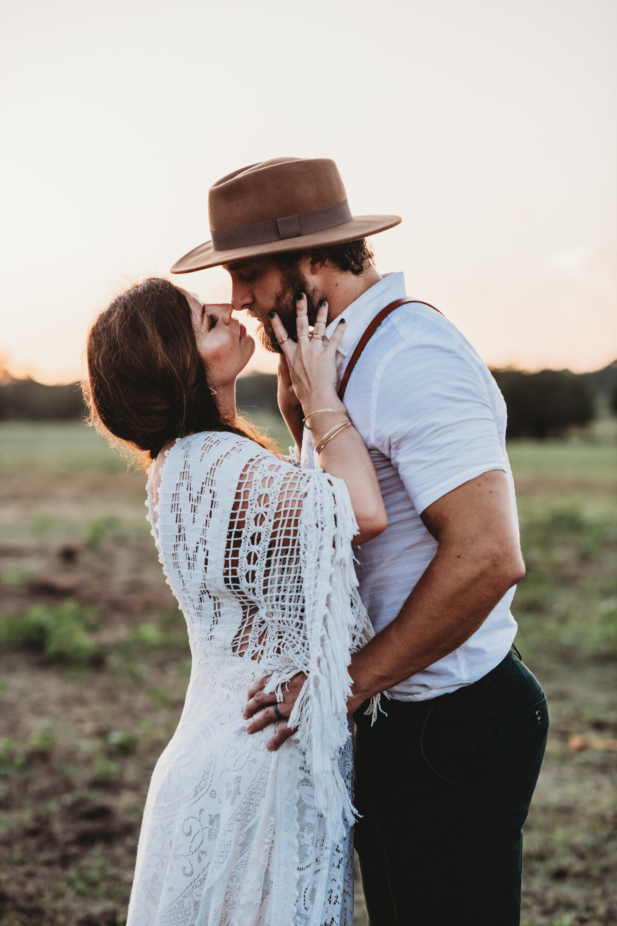 Couples Photography, woman in lacy white wedding dress kisses man in suspenders and hat in a dry meadow