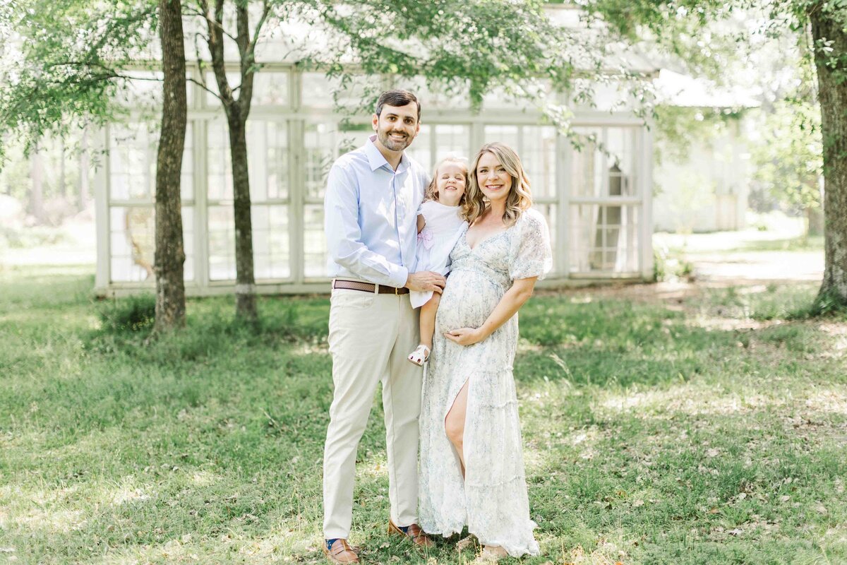 family of three smile together in front of conroe glass house