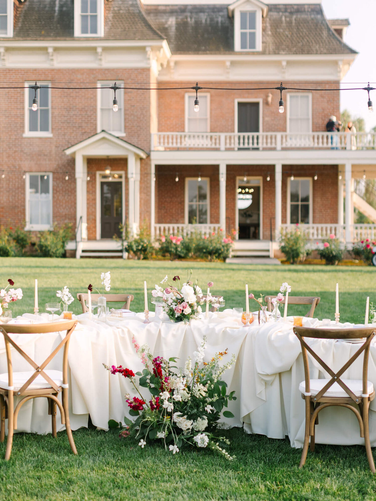Outdoor wedding setup of a long table adorned with white cloth, floral centerpieces, and candles in front of the Barton House