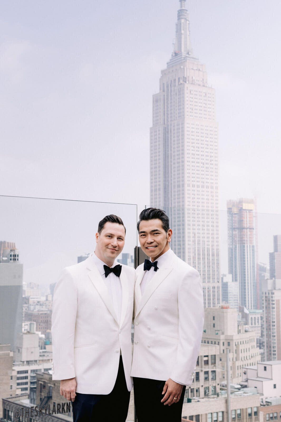 Two grooms are smiling, with buildings in the background, in The Skylark, New York. Wedding Image by Jenny Fu Studio