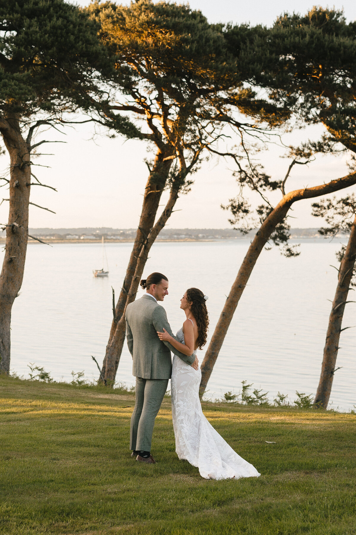 A bride and groom stand on a grassy lawn by the water, with tall trees framing the scene at their Hampshire wedding. The bride is in a white gown, and the groom is in a gray suit. A sailboat drifts by in the background, adding to the serene and picturesque setting.