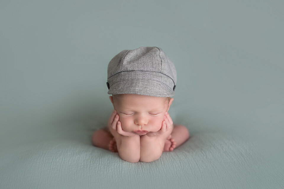 A newborn baby sleeps resting his head in his hands in a studio wearing a grey hat