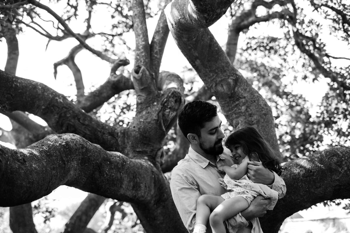 A father and daughter cuddle in front of a tree outdoors in a park in London