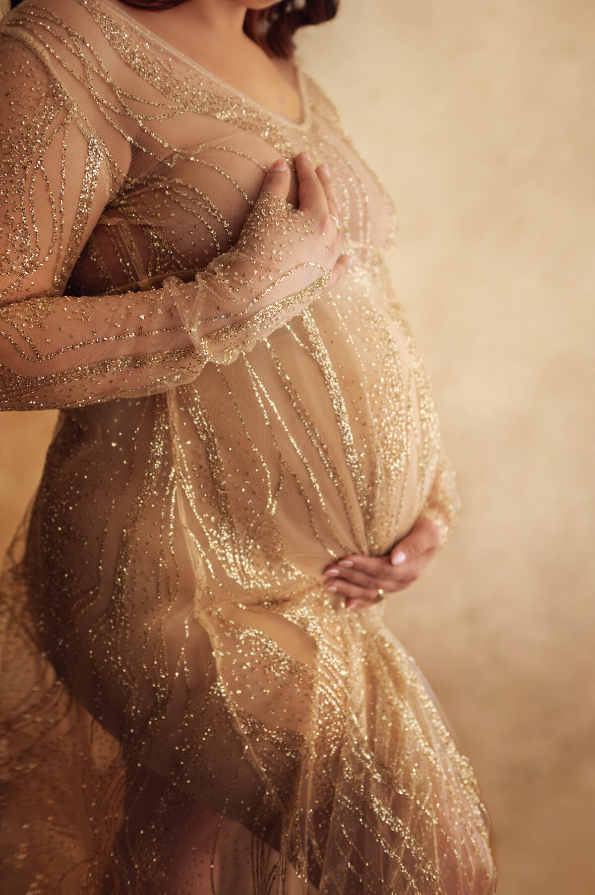 Close-up of a pregnant belly in a sparkling gold gown, hands gently cradling the bump in a glamorous and intimate maternity session near Pewaukee, Wisconsin.
