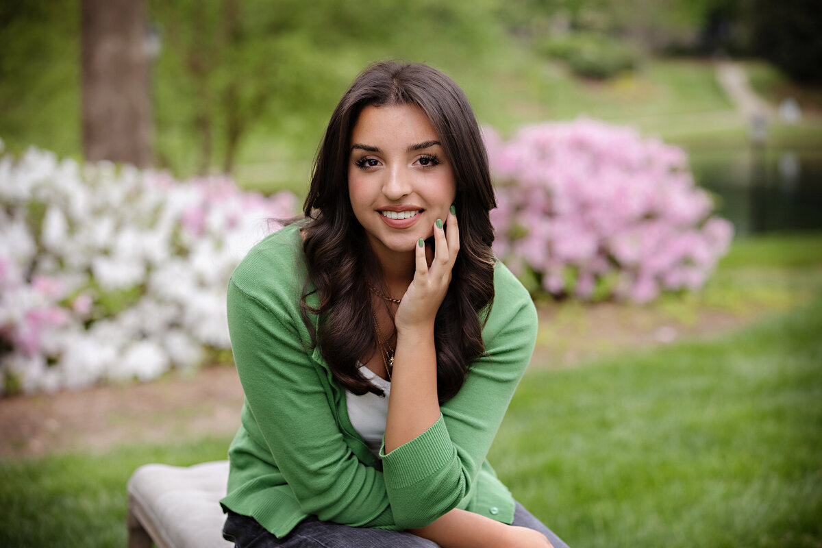 Smiling senior close-up with dark wavy hair and green cardigan, surrounded by blooming azaleas in Charlotte NC.
