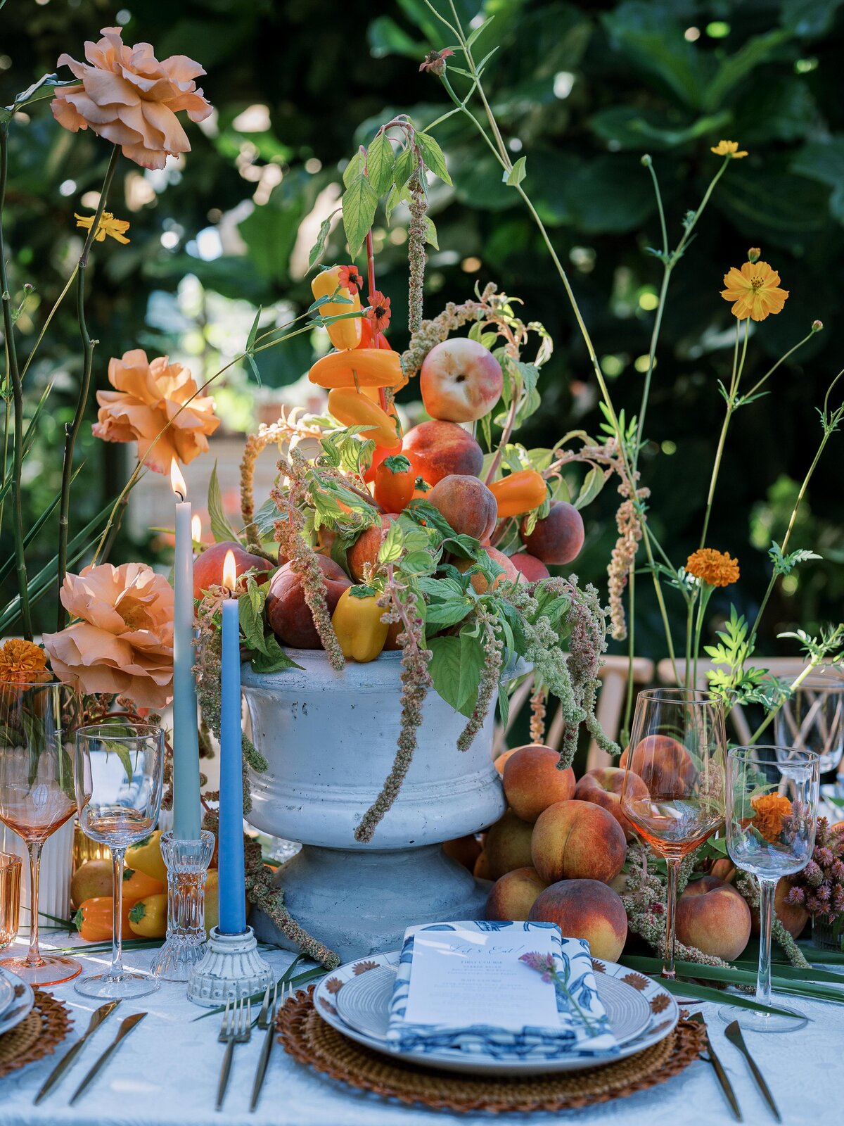 A lush outdoor tablescape featuring an abundant fruit and floral centerpiece with peaches, orange blooms, and cascading greenery. The setting includes elegant blue candles, woven placemats, and fine glassware, illuminated by natural sunlight.