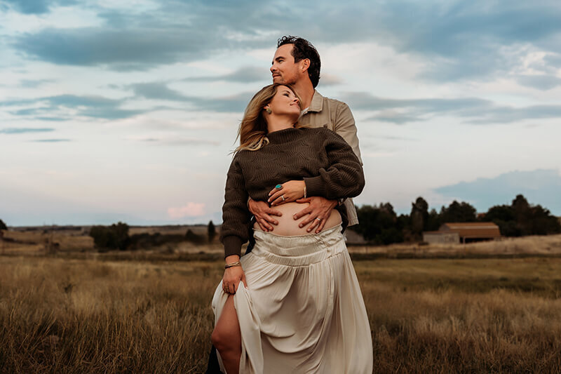husband embraces wife from behind while holding her exposed baby bump in a grassy field