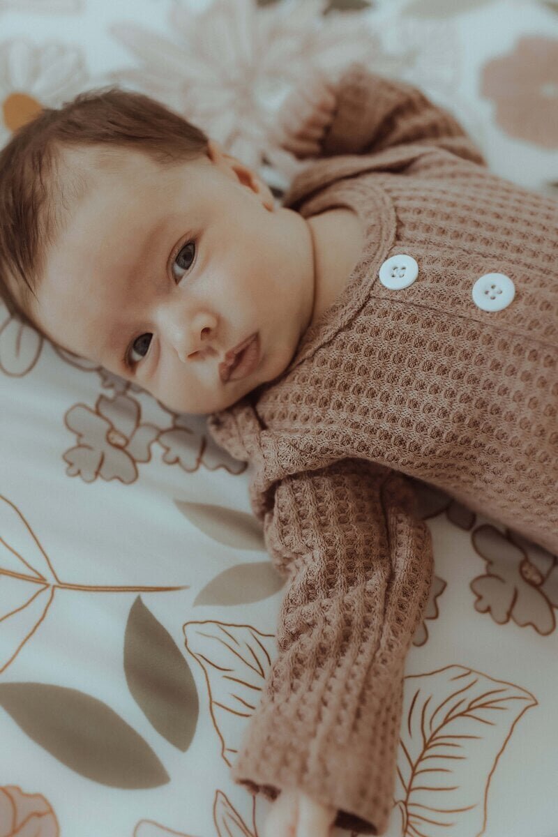 A baby lying on a patterned surface, wearing a textured brown outfit with two white buttons, looking at the camera.