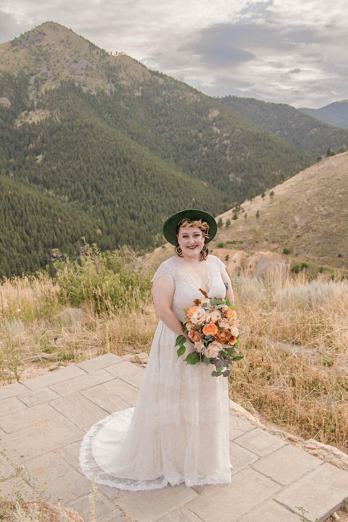 The bride, standing in front of a beautiful mountain covered in fir trees, holds her orange and pink bouquet while smiling for a photo. By Stephanie Eddy, Colorado wedding photographer.
