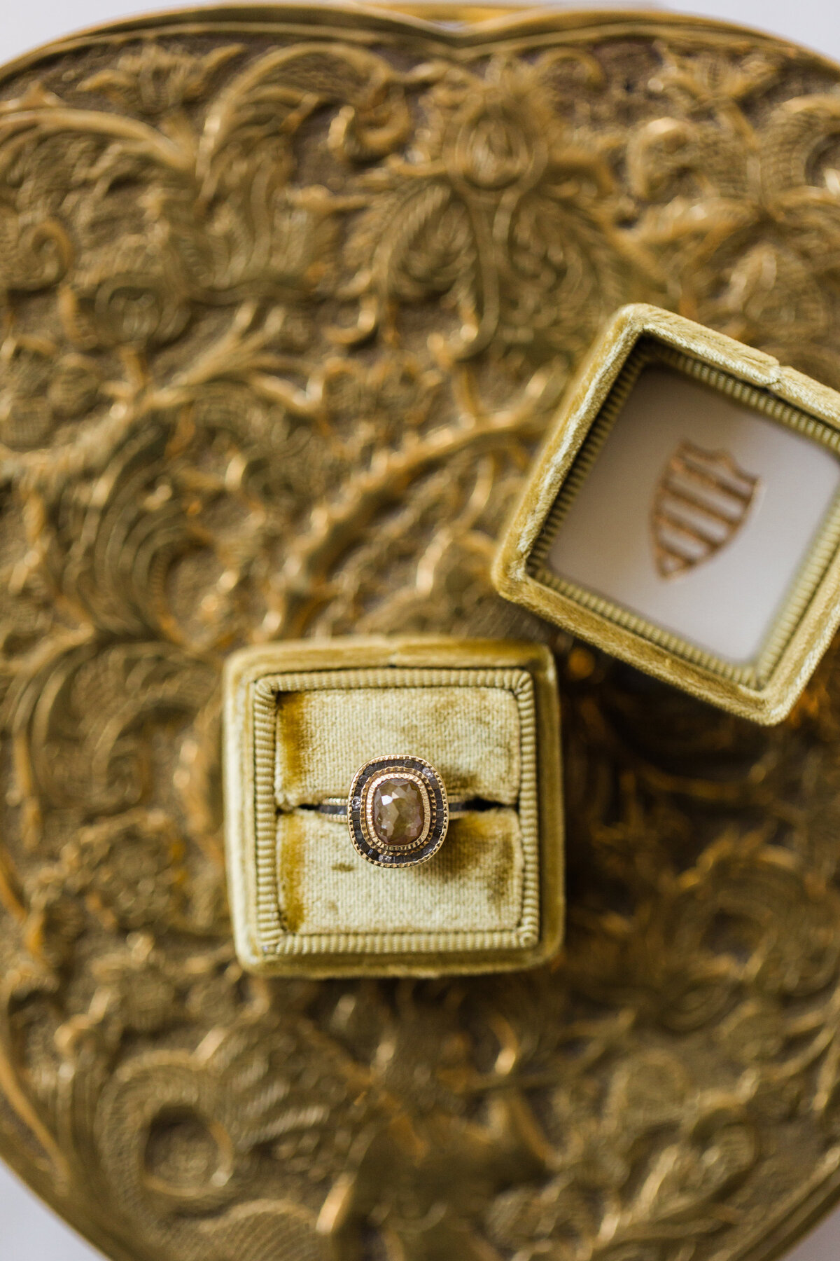 The stunning photo was taken at the Mellon Auditorium in Washington D.C., one of the best luxury venues in the city known for its European flair. Captured by the talented photographer Sarah Bradshaw, the photo showcases a close-up of a magnificent gold ring with a round gemstone set in a beautifully detailed, antique-style band. The ring is artfully presented in an elegant golden velvet ring box, with the lid slightly ajar to reveal a luxurious, ornate background that adds to its allure.