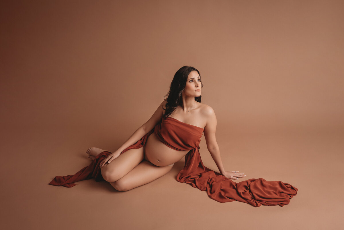 Pregnancy photography maternity portrait of woman sitting on floor with legs bent and looking over shoulder up to the right, draped in dark orange chiffon fabric