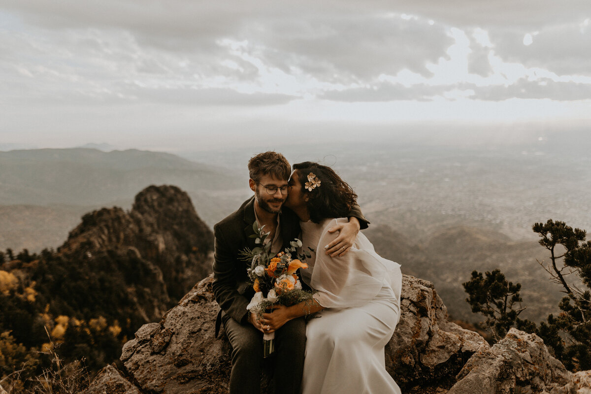 bride and groom sitting on the edge on the Sandia Mountain Peak with Albuquerque below them
