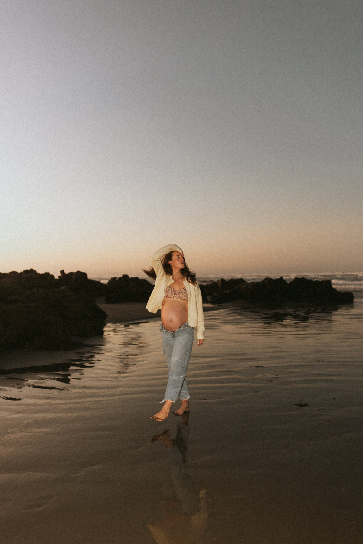 A woman wears a bra and loose oversized shirt and jeans and walks along the beach at blue hour during her maternity photoshoot in christchurch with lucy jane photography