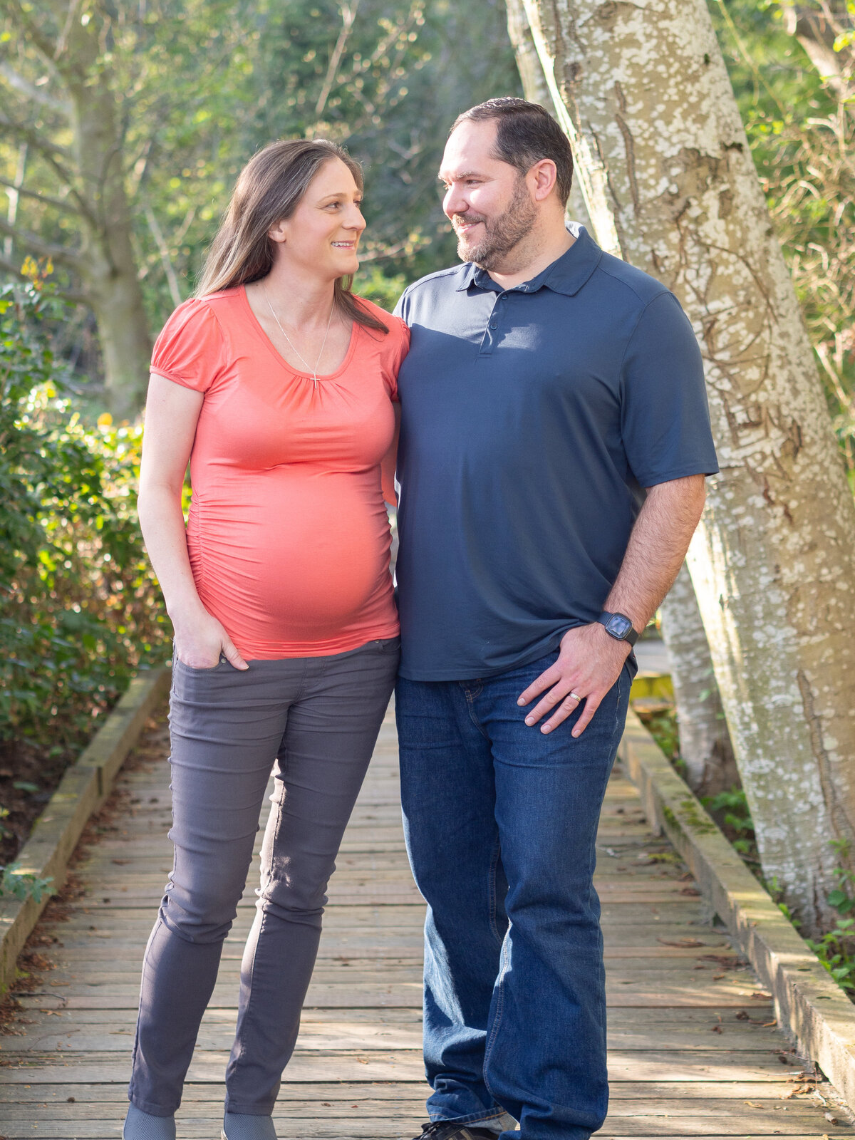 a pregnant couple look at each other as they stand long the Burke-Gilman trail in Lake Forest Park, WA.