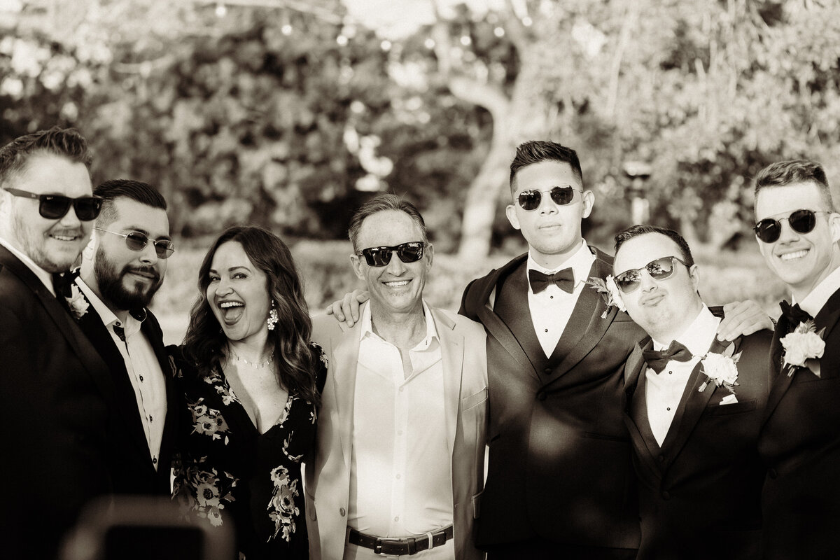groomsmen wearing sunglasses and black wedding tuxedos with bowties posing for a group photo during cocktail hour at spanish hills country club captured by los angeles wedding photographer magnolia west photography