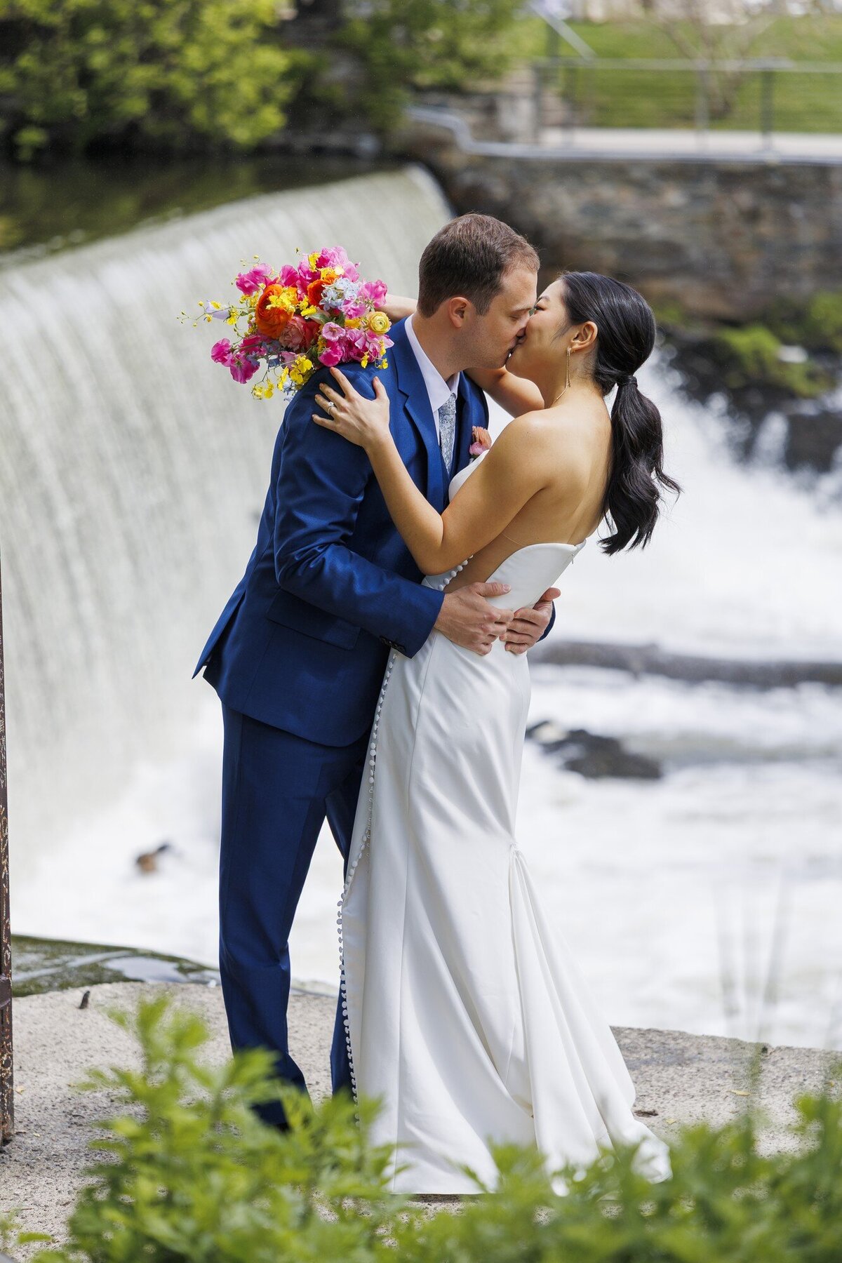 Couple sharing a joyful kiss in front of a waterfall with the bride holding a bright pink and orange bouquet