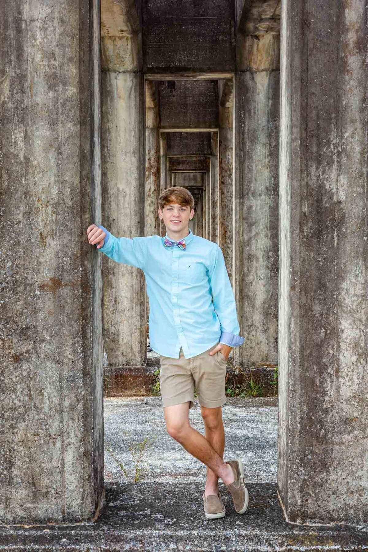 Senior portrait of a guy leaning against a cement column in the center of a row of columns.