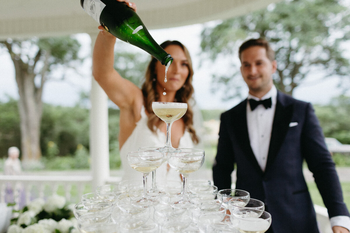 A woman pours champagne into a pyramid of glasses, with a man in a suit standing beside her at their Brighthouse Farm wedding. Captured by Jennifer van Son, a destination photographer from Toronto, they are outdoors surrounded by lush greenery.
