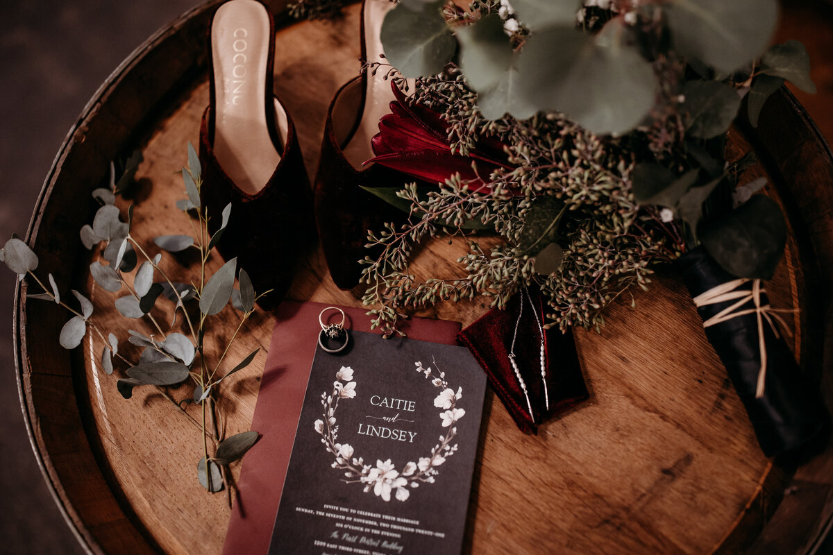 Close-up of a wedding invitation, a pair of high heels, and a bridal bouquet arranged on a wooden barrel