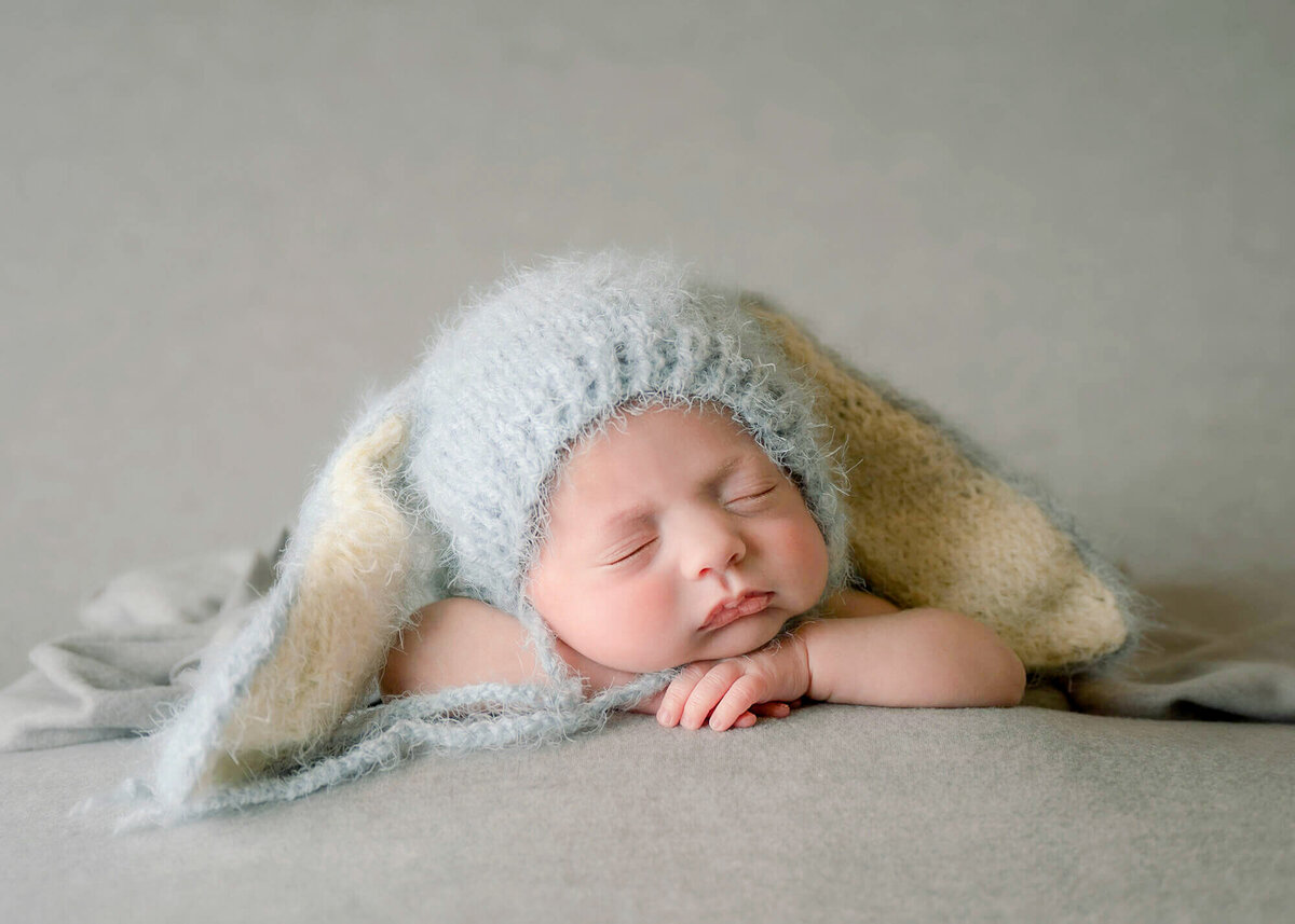 Small baby laying on his hands  and wearing a blue  rabbit ear hat.