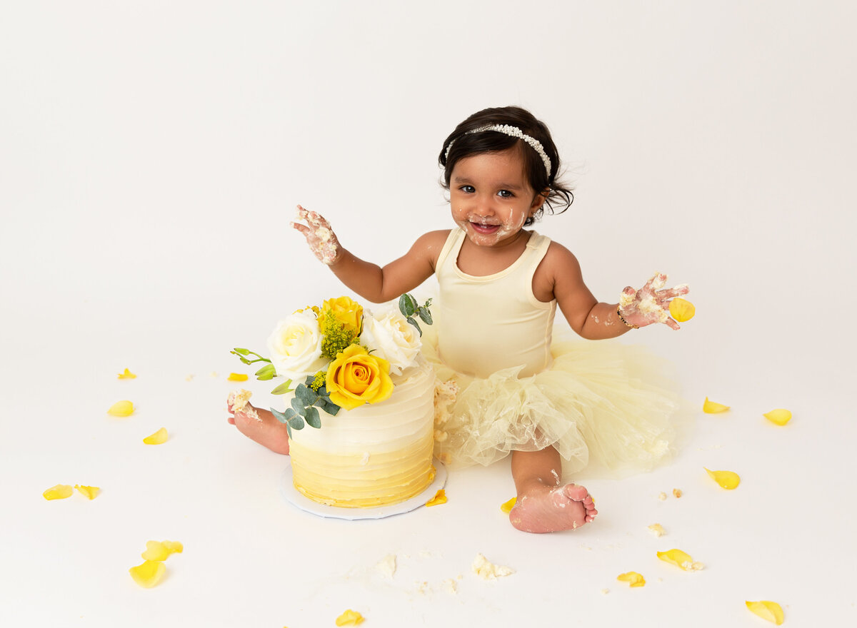 Baby girl in yellow tutu sits with yellow ombre cake topped with yellow roses between her legs. She has icing on her hands and face.