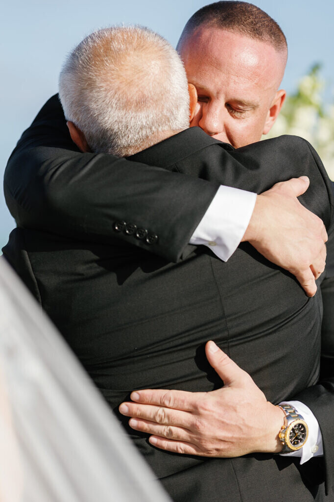 An emotional moment as the bride's father embraces the groom before entrusting his daughter to him at their oceanfront wedding ceremony. Destination wedding photography by Claudia Amalia Photography.