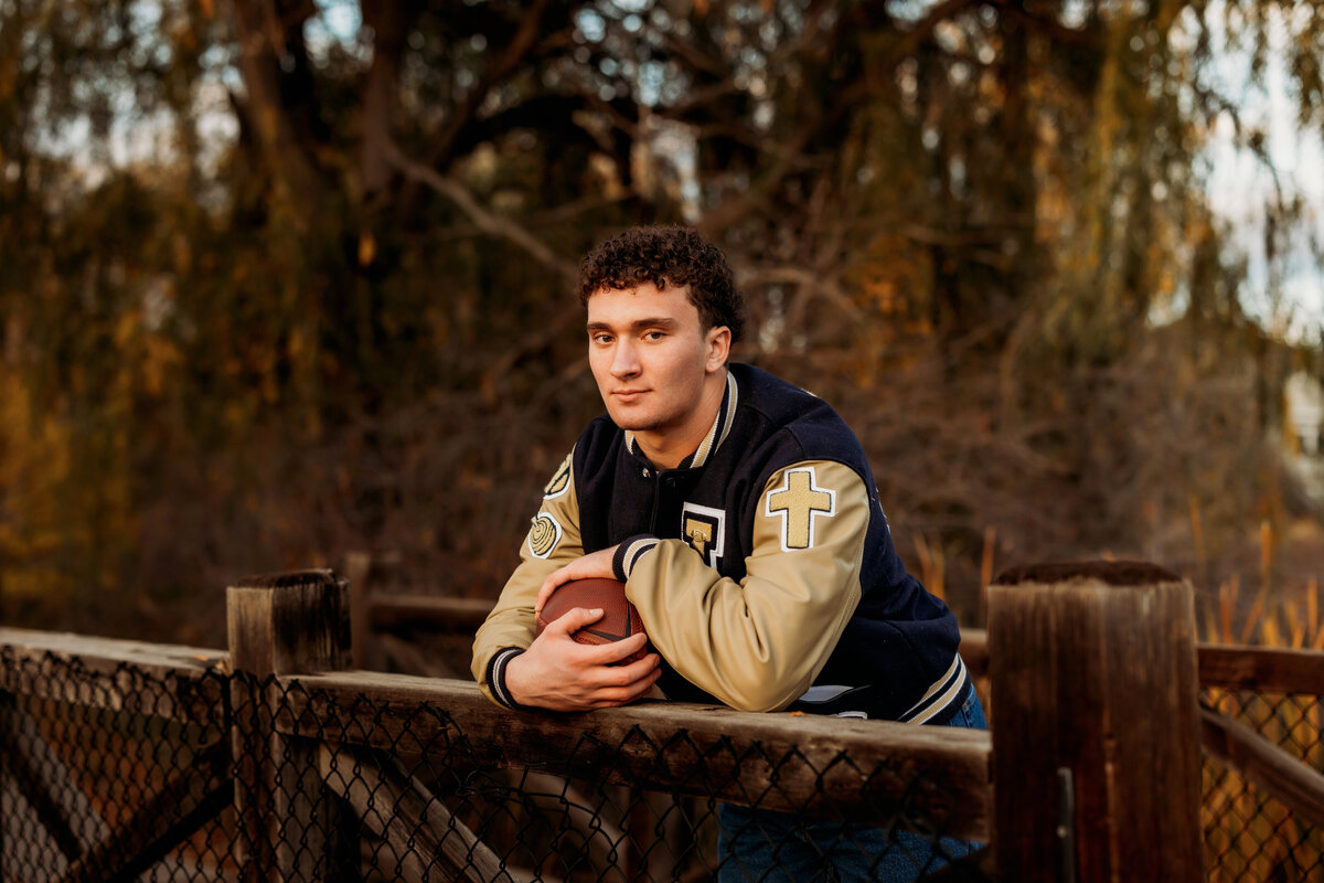 Senior boy leaning over a wooden bridge while wearing a letterman's jacket and holding a football for his high school senior photos