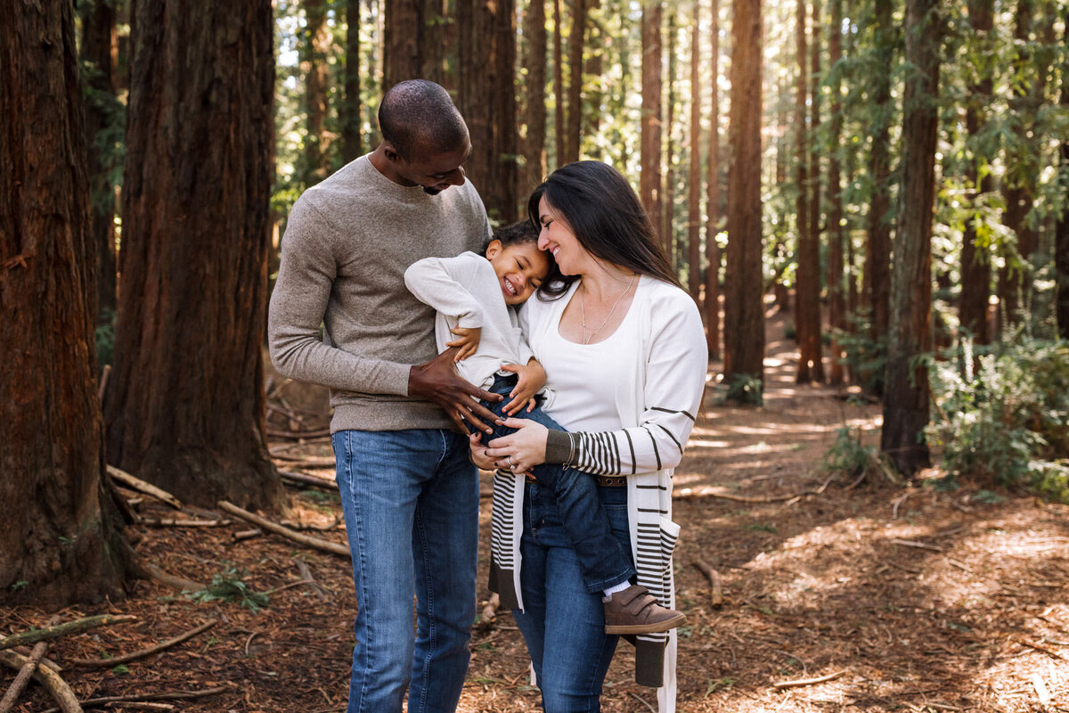 mom and dad holding son in Oakland woods with him looking towards camera  and laughing while they look at him and smile