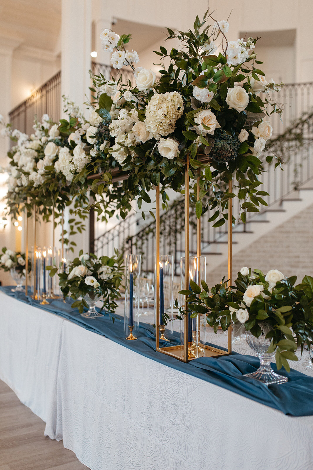 White floral arrangements in gold lanterns above a long rectangular table with blue and white linen.