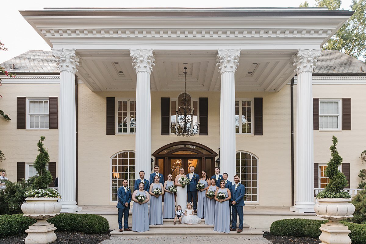 Groomsmen dressed in dark blue suits and bridesmaids dressed in light blue dresses holding white bouquets of roses and hydrangea made my a Nashville Florist pose with the bride and groom in front of the mansion at The Estate at Cherokee Dock.