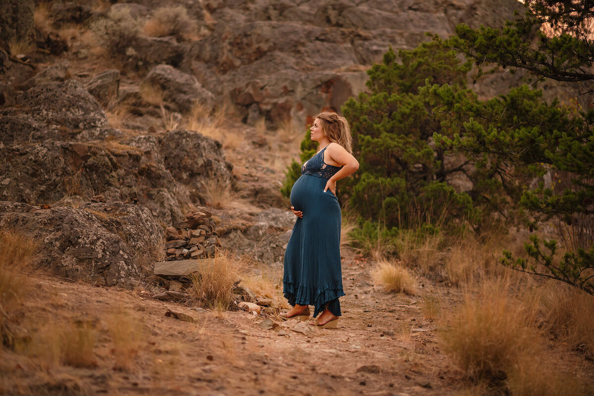 A pregnant women wearing a teal dress stands looking to the left while caressing her belly. She is looking off into the distance and there is a rocky terrain behind her. Captured in Kamloops by Brittany Danielle Photography
