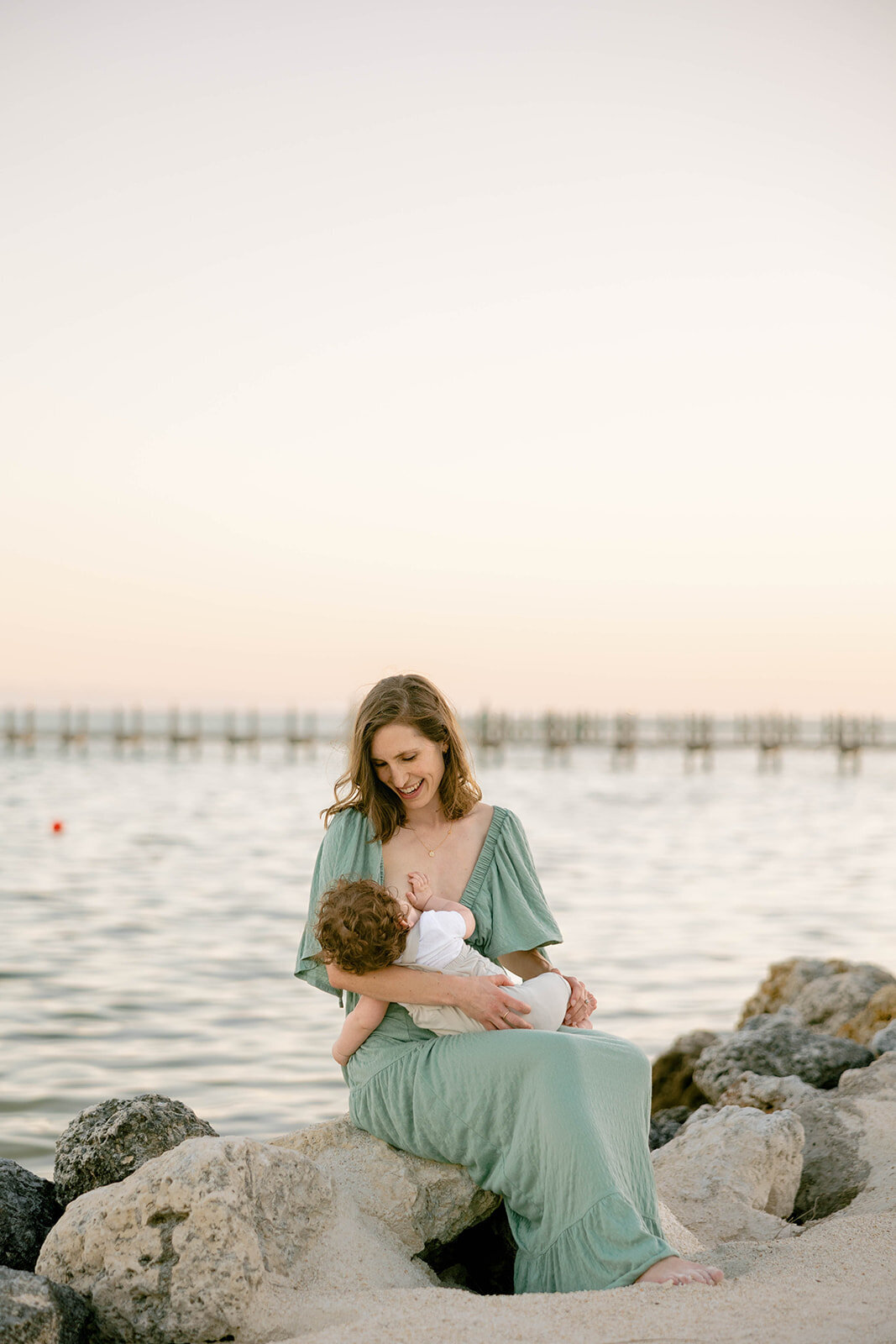 A mother and child share a tender moment sitting on rocks by the shore during a Florida Keys portrait session. Claudia Amalia Portrait Photography.