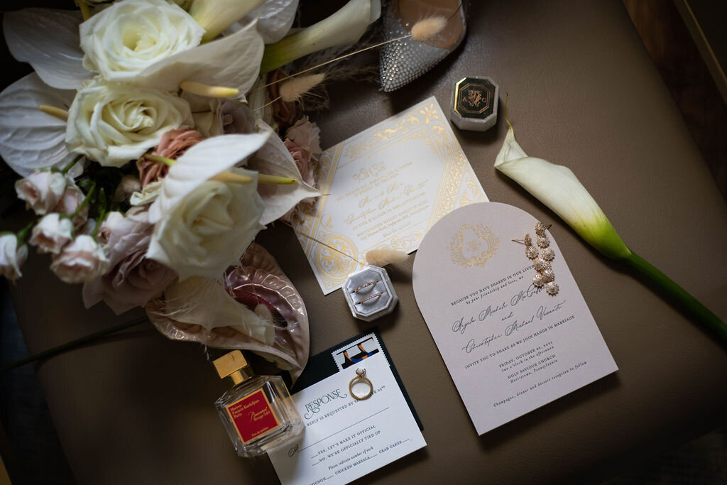 A flat lay of wedding essentials, including an elegant invitation, vow books, perfume, and a delicate floral arrangement.