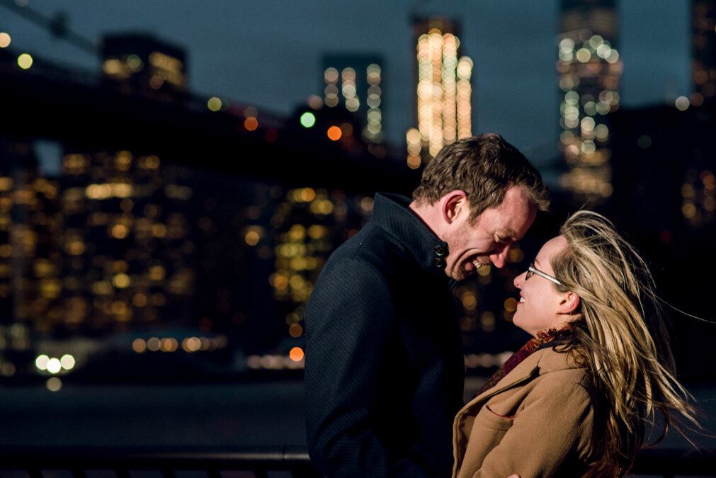 A couple embracing in front of the brooklyn bridge at night.