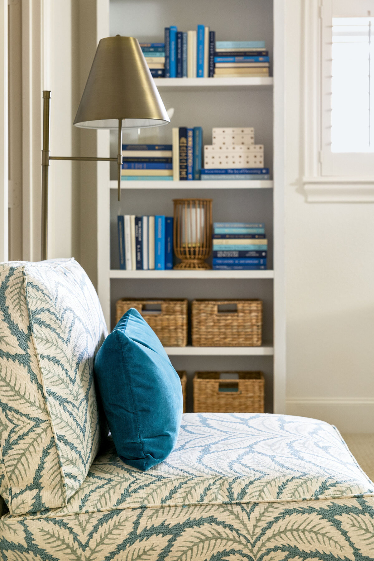 Profile shot of lounge chair with blue leaf pattern fabric  and velvet accent pillow. White bookcase accessorized with blue books, rattan hurricanes, and woven baskets