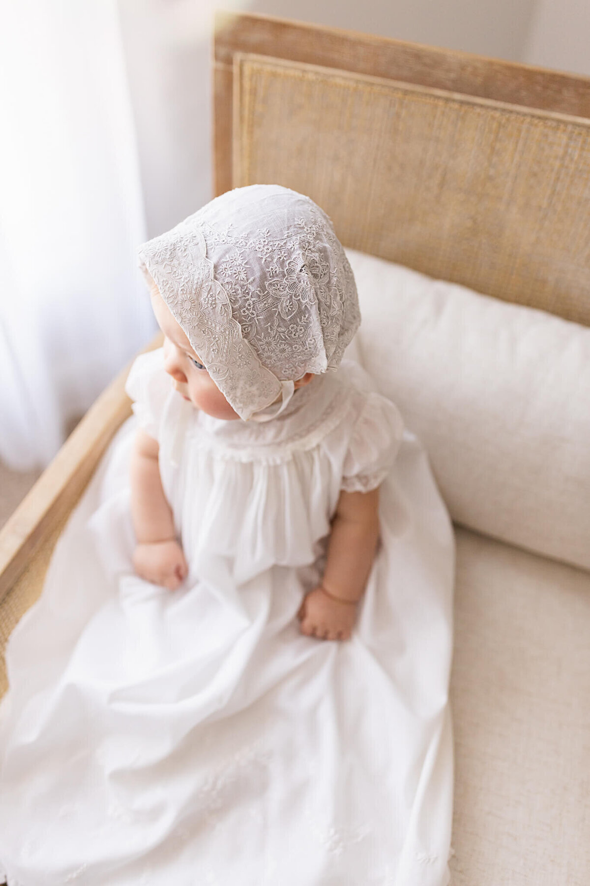 Baby girl sits in a chair wearing a white dress and bonnet