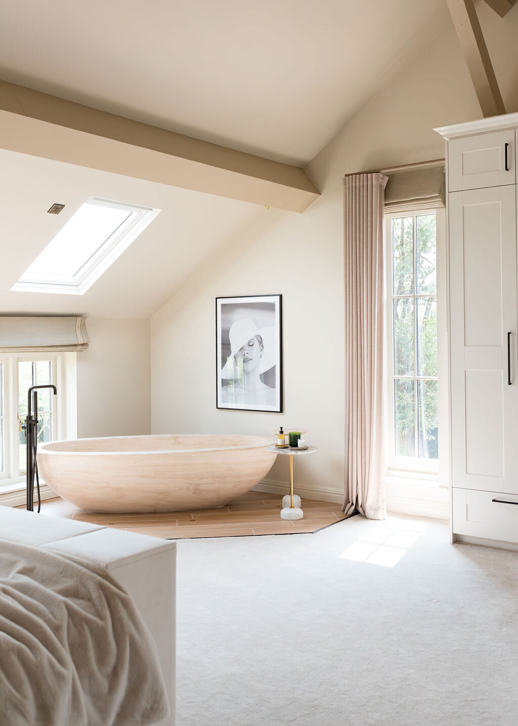 A serene bedroom with a freestanding wooden bathtub on a wooden platform beneath a slanted ceiling with a skylight. There's a window with sheer curtains to the right, a framed black-and-white portrait on the wall, and minimalist decor throughout.