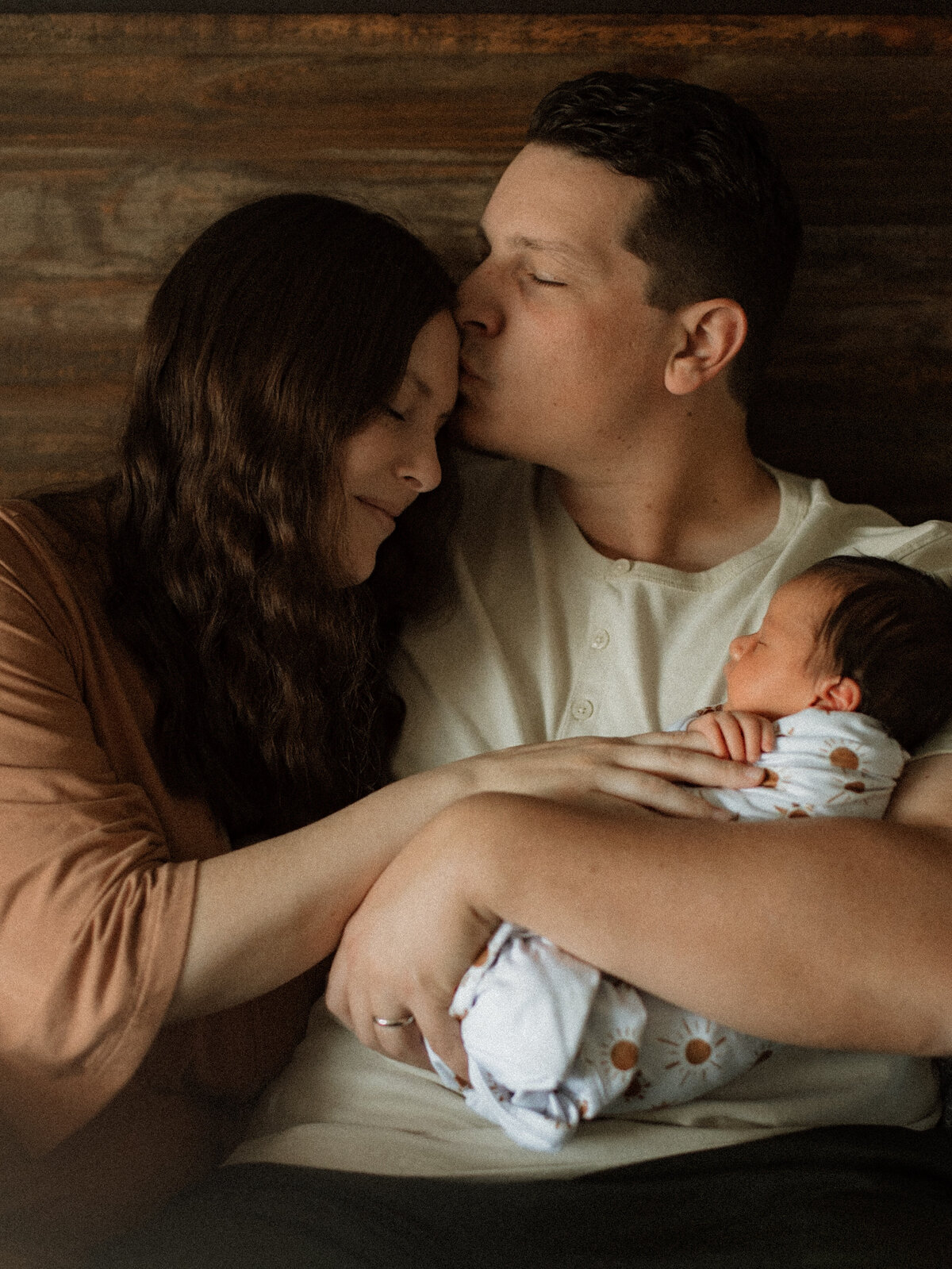 Parents embracing their swaddled newborn on a bed, forehead kiss and soft expressions of love.
