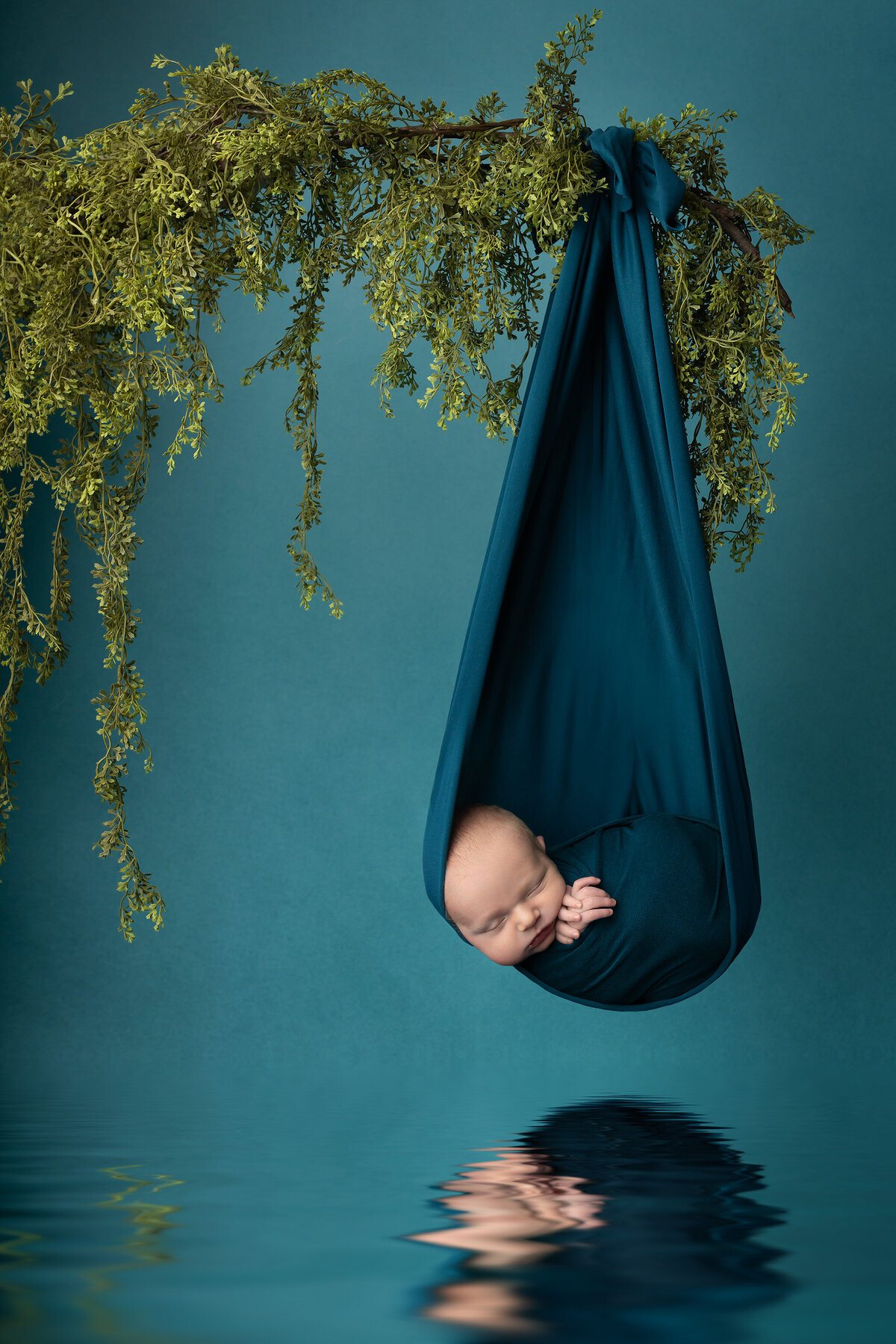 Newborn baby wrapped in teal fabric hanging from a branch with greenery, peacefully sleeping above a water reflection, captured in a fine art Dallas newborn photography session.