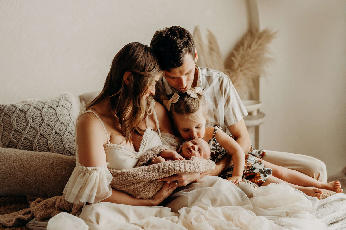 toddler sister kisses her baby brother's head as his mom and dad cuddle close