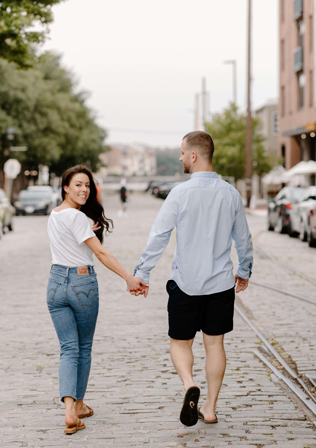 couple holding hands and looking back on cobblestone city road in baltimore maryland by sabrina leigh photography